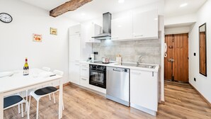Eine moderne Küche mit weißen Schränken, einem eingebauten Backofen und einem Geschirrspüler. Ein Holzbalken in der Decke, eine Uhr an der Wand und eine kleine Essecke mit Tisch und Stühlen.