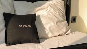 Cama com roupa de cama branca e almofada preta com a inscrição 'DS COLIVE'.