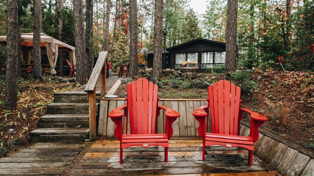 Une terrasse en bois avec deux chaises Adirondack rouges, un escalier en bois et un chalet situé dans une zone boisée.