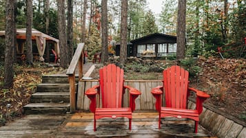 A wooden deck with two red Adirondack chairs, a wooden staircase, and a cabin in a forested area.