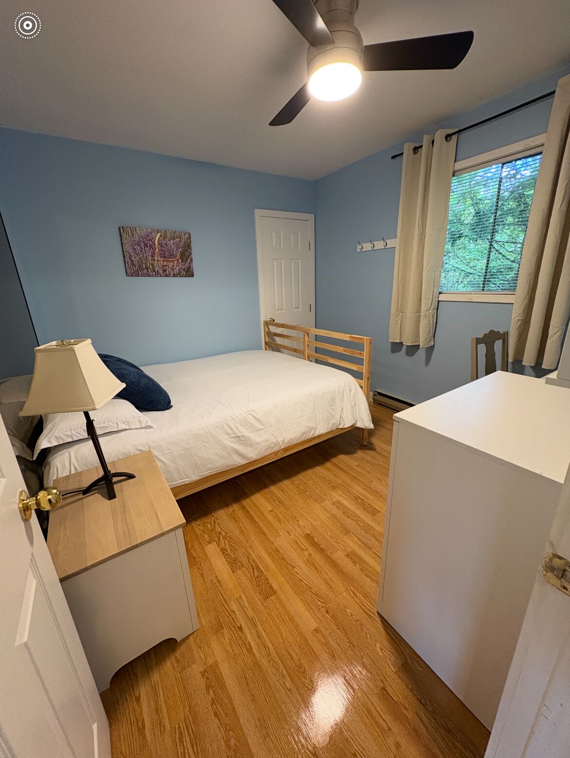 A bedroom with a bed, a nightstand, a ceiling fan, a window with curtains, and a door.