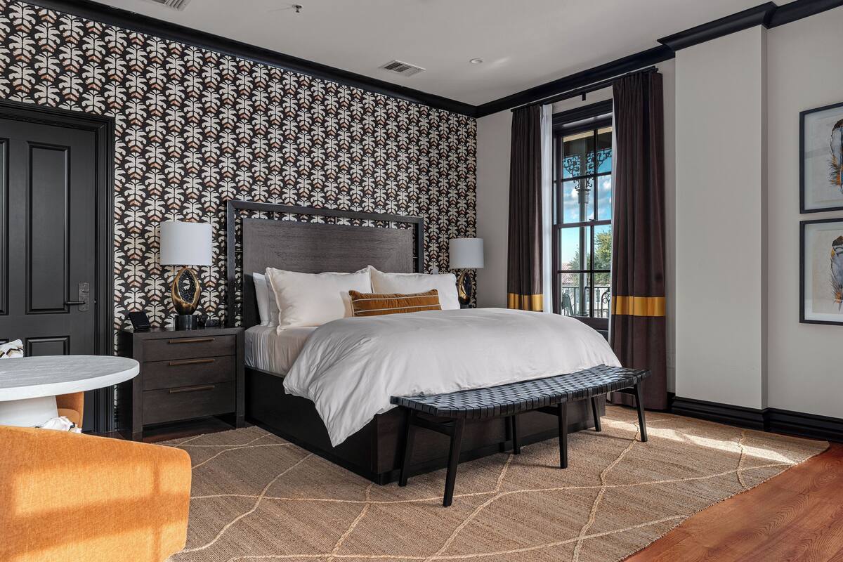 A hotel room with a large bed, bedside lamps, a round table, a bench, and a patterned wallpaper.