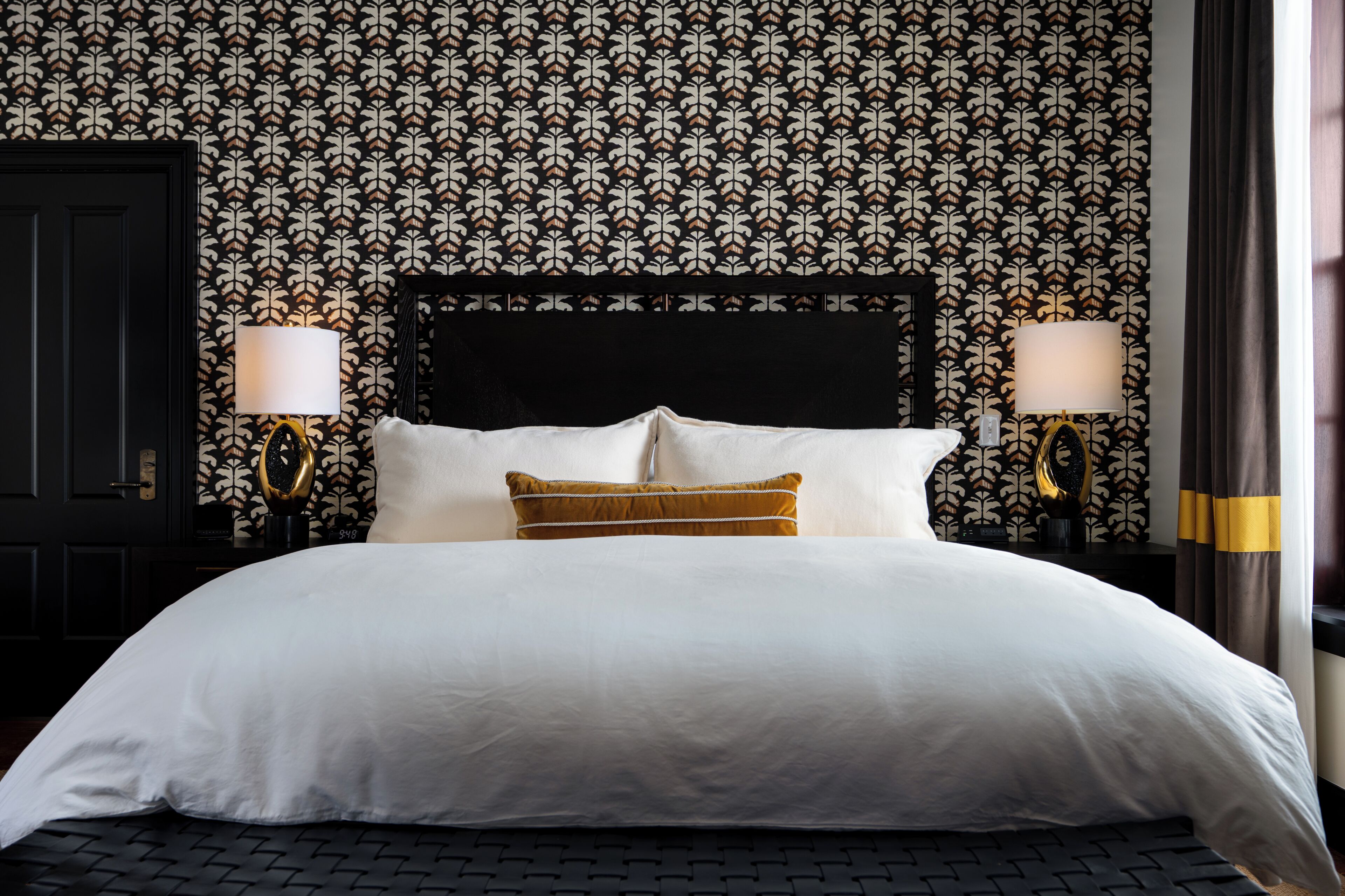 A hotel room with a patterned wallpaper, a black headboard, two bedside lamps, and a bed with white and mustard-colored bedding.