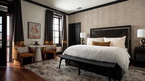 Une chambre avec un grand lit, deux tables de chevet avec des lampes, un banc, une petite table avec une chaise et une fenêtre avec des rideaux.