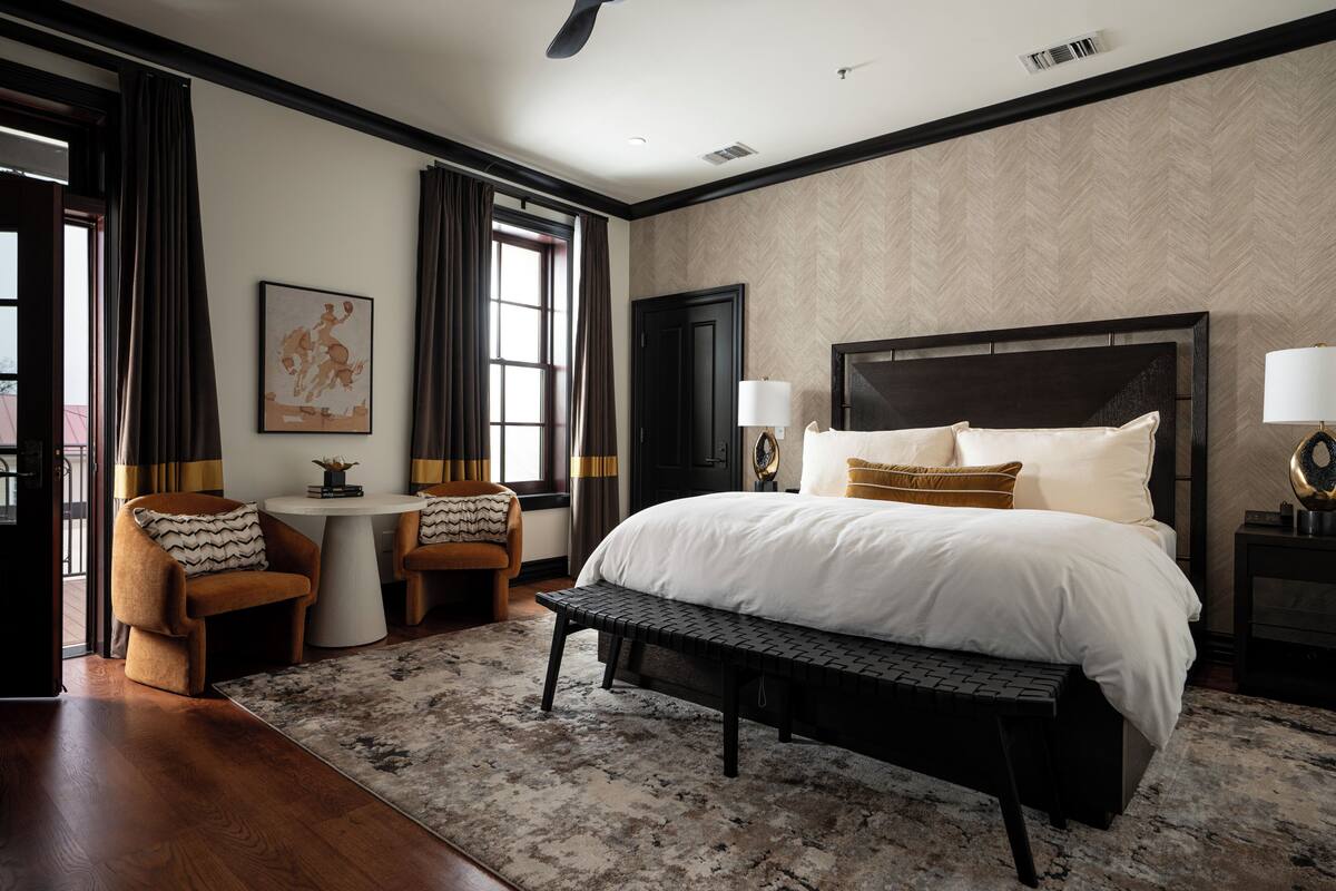A bedroom with a large bed, two bedside tables with lamps, a bench, a small table with a chair, and a window with curtains.