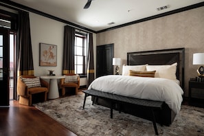 Un dormitorio con una cama grande, dos mesitas de noche con lámparas, un banco, una pequeña mesa con una silla y una ventana con cortinas.