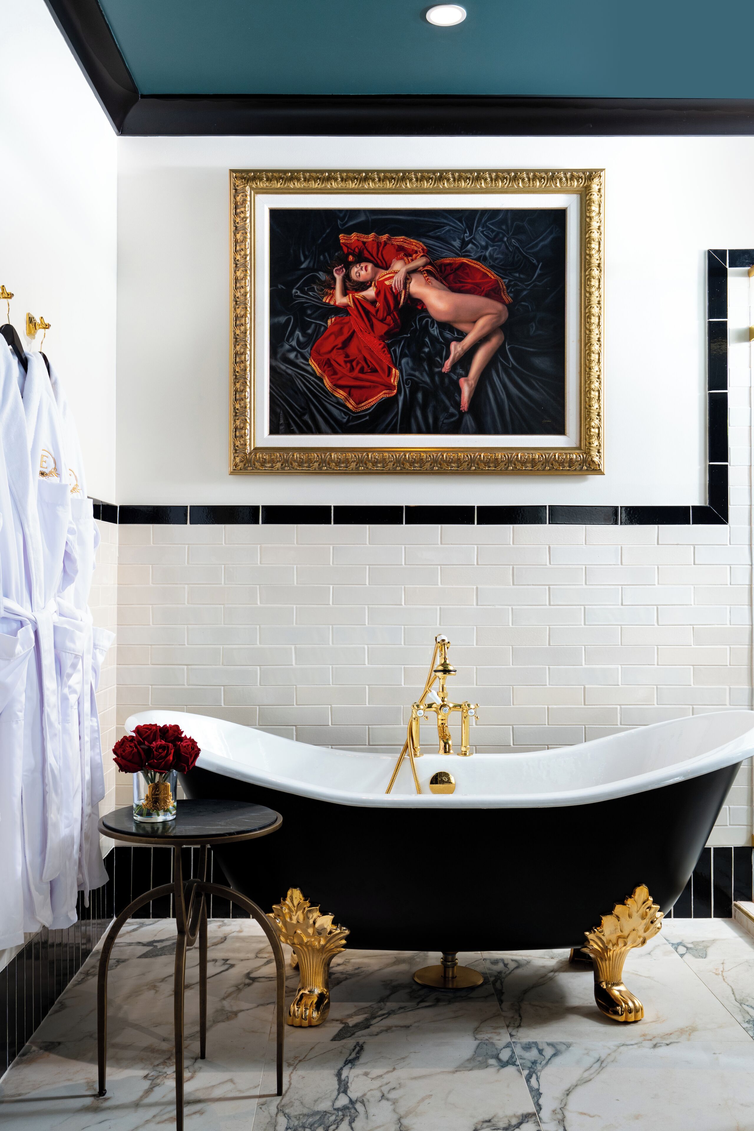 A bathroom with a black bathtub, white tiled walls, a gold-framed painting, and a small table with a vase of red flowers.
