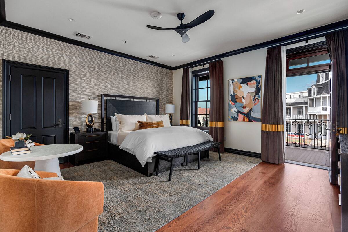 A hotel room with a bed, a desk, a chair, a small table, a ceiling fan, and a large window with curtains.