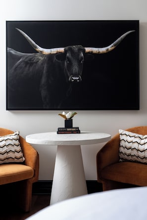 A framed artwork of a bull's head on a wall, a small round table with books, and two brown armchairs with patterned cushions.