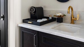 Une salle de bain avec un meuble-lavabo en bois foncé, un évier blanc, un robinet doré, une machine à café noire, un miroir fixé au mur et une petite lampe murale.