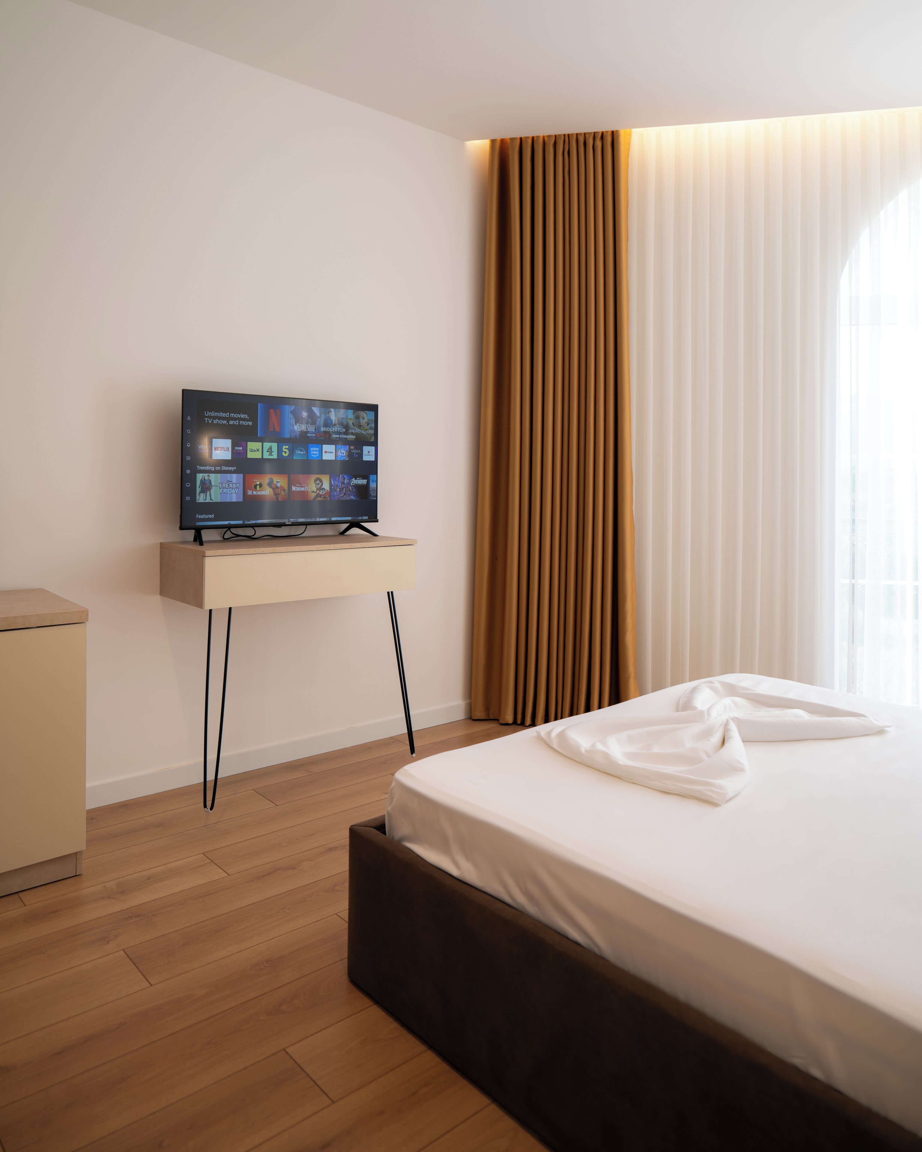 A hotel room with a bed, a TV on a stand, a nightstand, and a window with curtains.