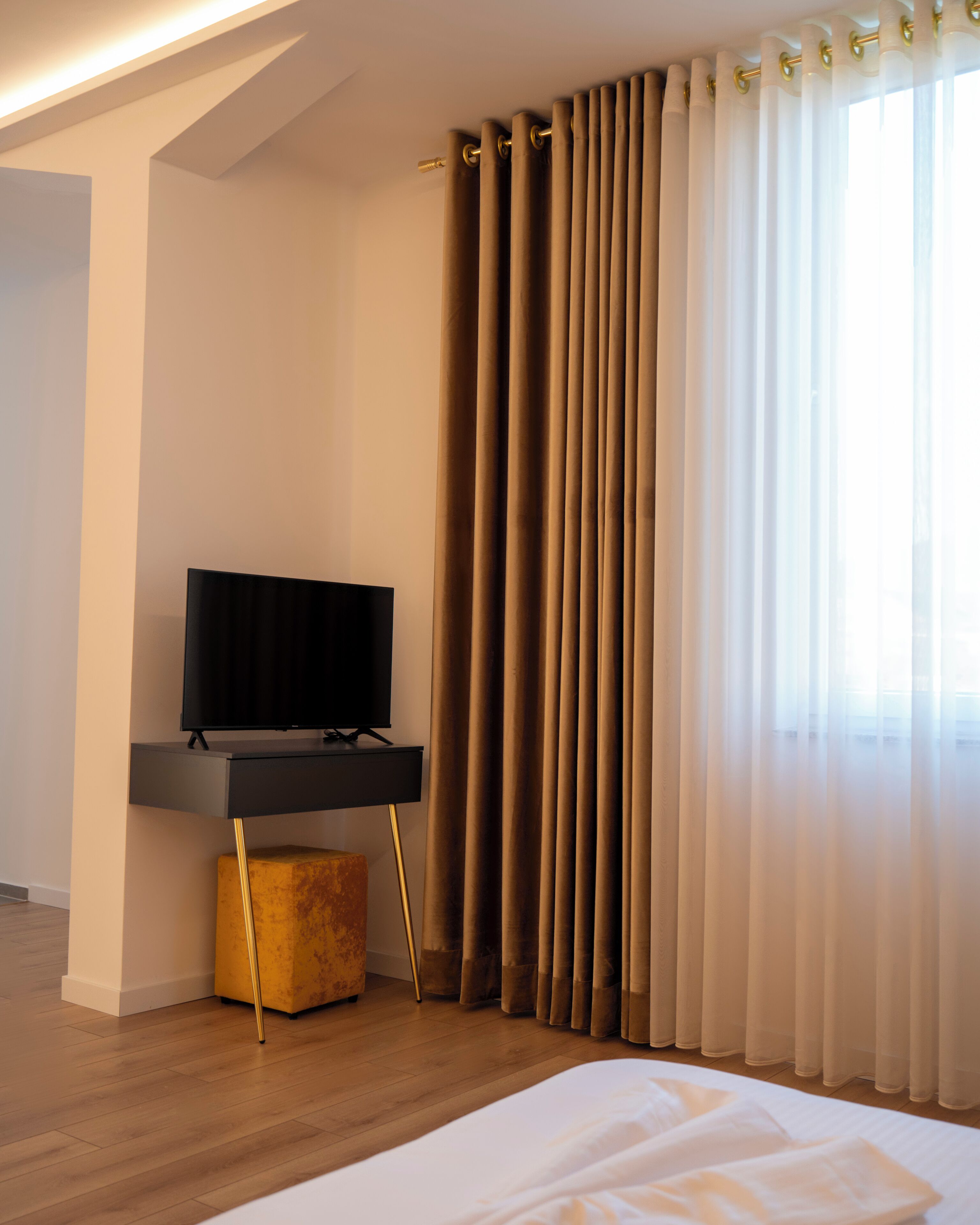 A modern hotel room with a flat-screen TV on a black stand, a wooden floor, a bed with white linens, and a large window with sheer curtains.