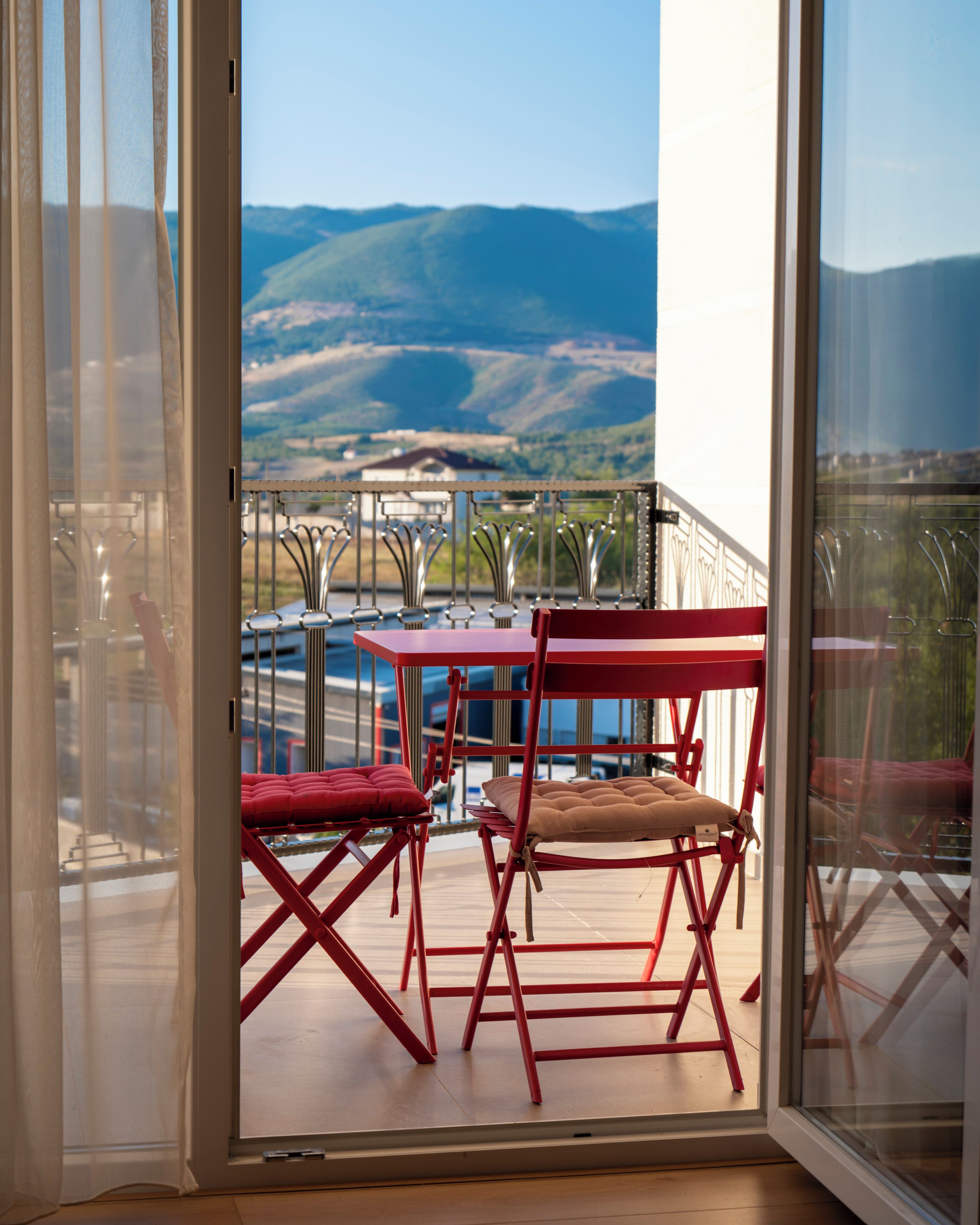 A balcony with a red metal table and chairs, a glass door, and a view of mountains and a body of water.