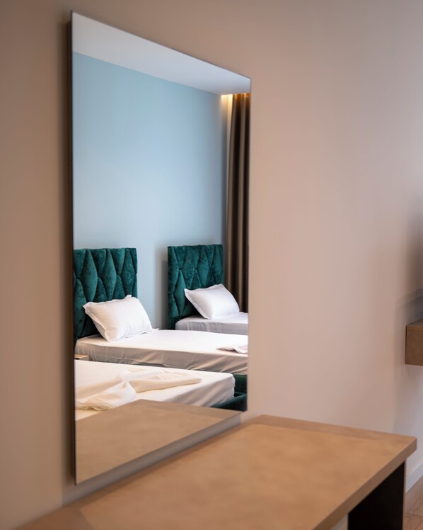 A hotel room with two beds, each with a teal headboard and white bedding. A large mirror reflects the room, and a wooden bench is in the foreground. - Leo Hotel (Shtiqën)