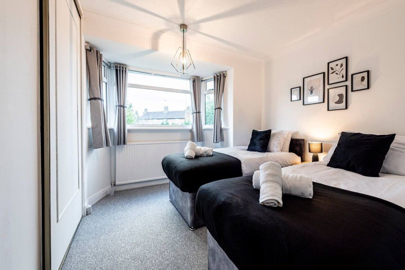 Une chambre d’hôtel avec deux lits, une fenêtre avec des rideaux, un mur décoré de tableaux encadrés et un luminaire au plafond.
