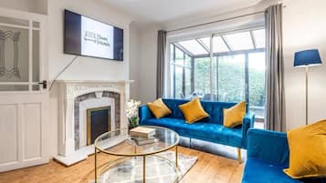 A living room with a blue sofa, a fireplace, a television mounted on the wall, and a large window offering a view of greenery.