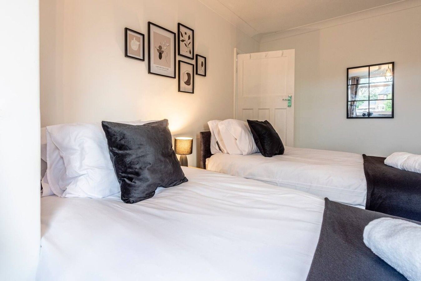 Ein Hotelzimmer mit zwei Betten, weißer Bettwäsche, schwarz-weißen Kissen, einem Nachttisch mit Lampe und gerahmten Kunstwerken an der Wand.
