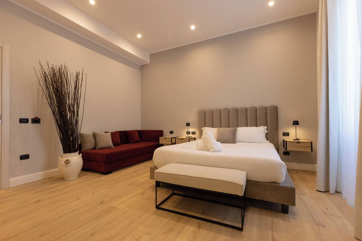 A modern bedroom with a large bed, a red sofa, a bedside table with a lamp, and a tall vase with branches.