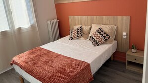 A neatly made bed with a patterned throw blanket, a bedside table with a plant, and a wall-mounted light fixture. - Central Hotel (Saint-Priest)