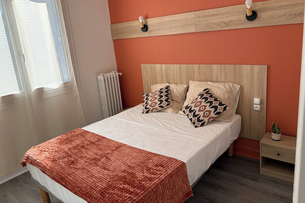 A neatly made bed with a patterned throw blanket, a bedside table with a plant, and a wall-mounted light fixture.