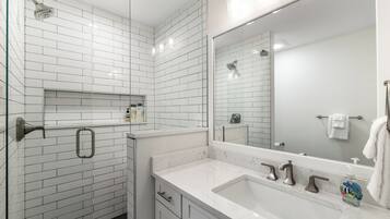 Ein modernes Badezimmer mit weißen, rechteckigen Fliesen, einem großen Spiegel, einer Dusche und einem Waschtisch mit Spüle und Handtuchhalter.