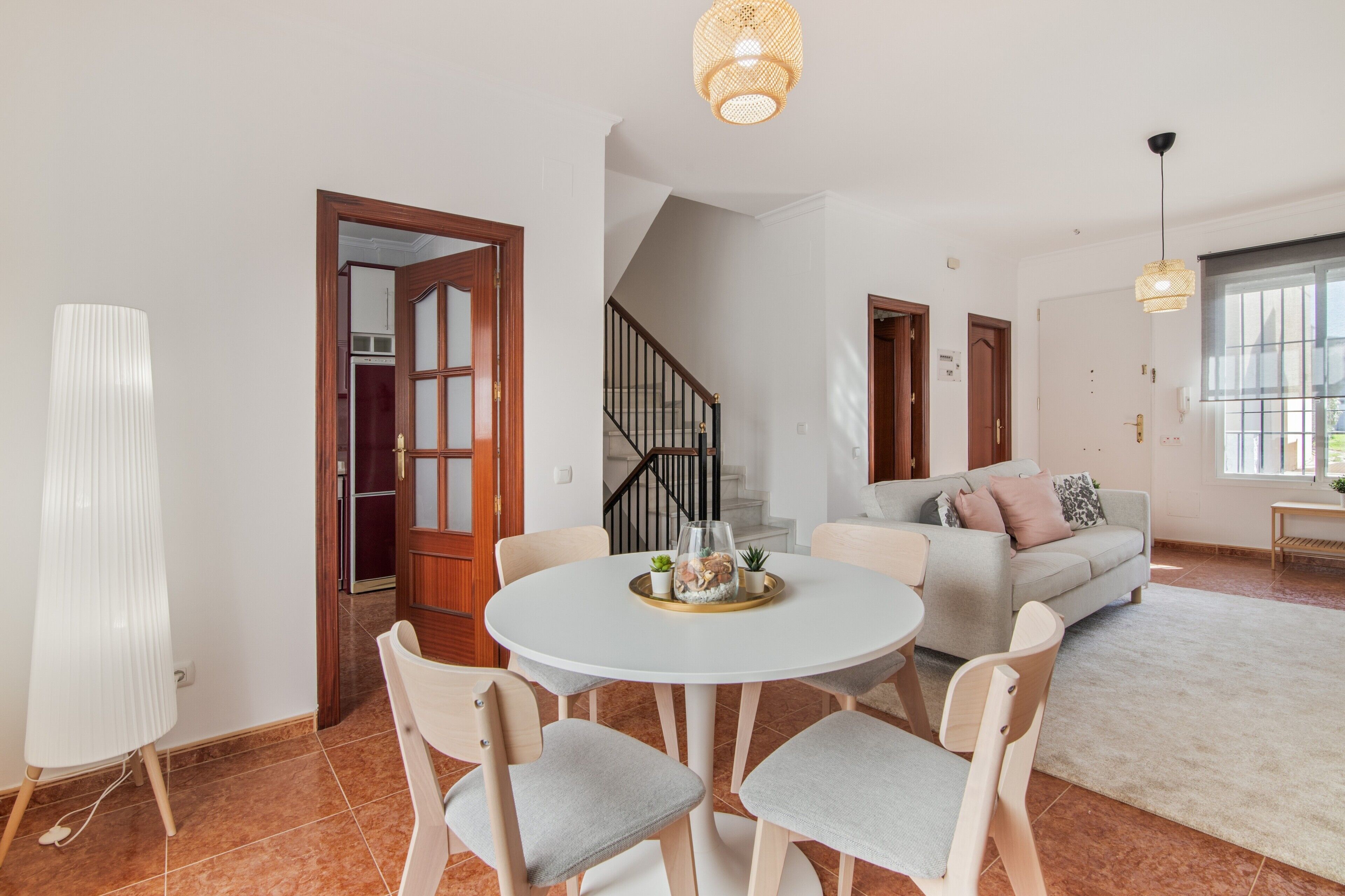 Un vaste espace de vie comprenant une table à manger ronde, des canapés aux couleurs claires, un escalier et une vue sur l’extérieur.