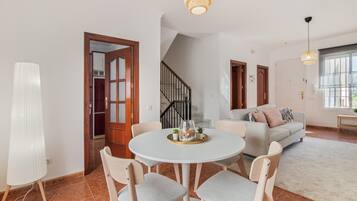 Un vaste espace de vie comprenant une table à manger ronde, des canapés aux couleurs claires, un escalier et une vue sur l’extérieur.