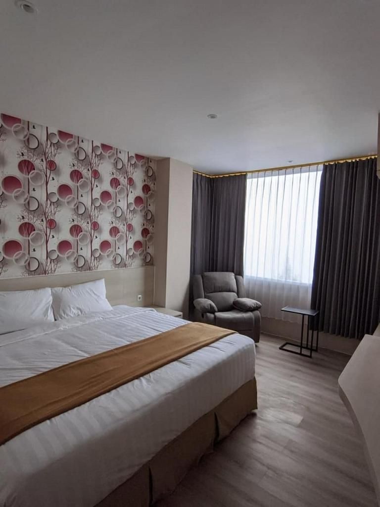 A hotel room with a large bed, a chair, a small table, and a window with curtains.