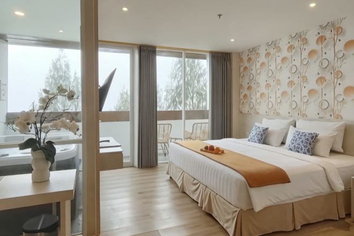 Una habitación de hotel moderna con una cama grande, un rincón de estar con una mesa y sillas, vistas a través del ventanal y un detalle decorativo en la pared.