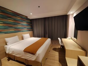 Quarto de hotel com cama, escrivaninha, cadeira, televisão e um quadro na parede.