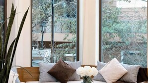 A room with a sofa, pillows, a vase with flowers, and large windows with a view of trees and buildings. - Adalia Villa With ValeyView - O'TEL (Dalat)