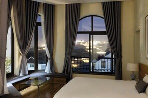 A hotel room with a large bed, a view of a cityscape through a window, and a balcony with a railing. - Adalia Villa With ValeyView - O'TEL (Dalat)