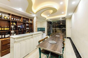Un espace bar avec une longue table en bois et des chaises, un comptoir de bar avec des étagères remplies de bouteilles, et un plafond au design incurvé.