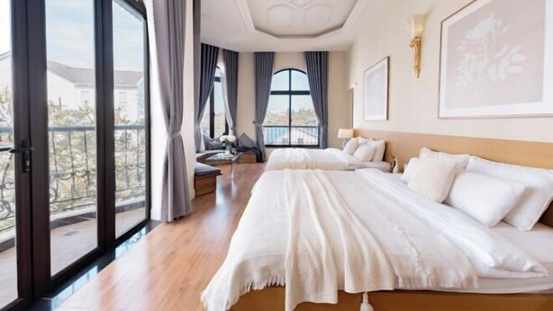 A hotel room with a large bed, a sofa, and a view of the city through large windows. - Adalia Villa With ValeyView - O'TEL (Dalat)