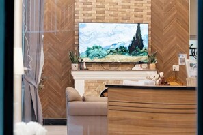 A reception area with a flat-screen TV mounted on a brick wall, a sofa, and a wooden reception desk. - Adalia Villa With ValeyView - O'TEL (Dalat)