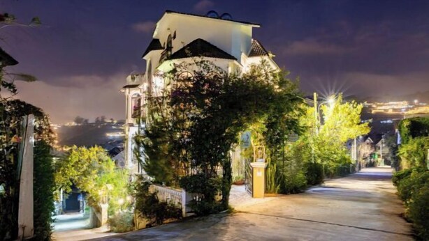 Un chemin bien éclairé mène à un bâtiment à la façade blanche et au toit foncé, entouré de verdure et sous un ciel dégagé.