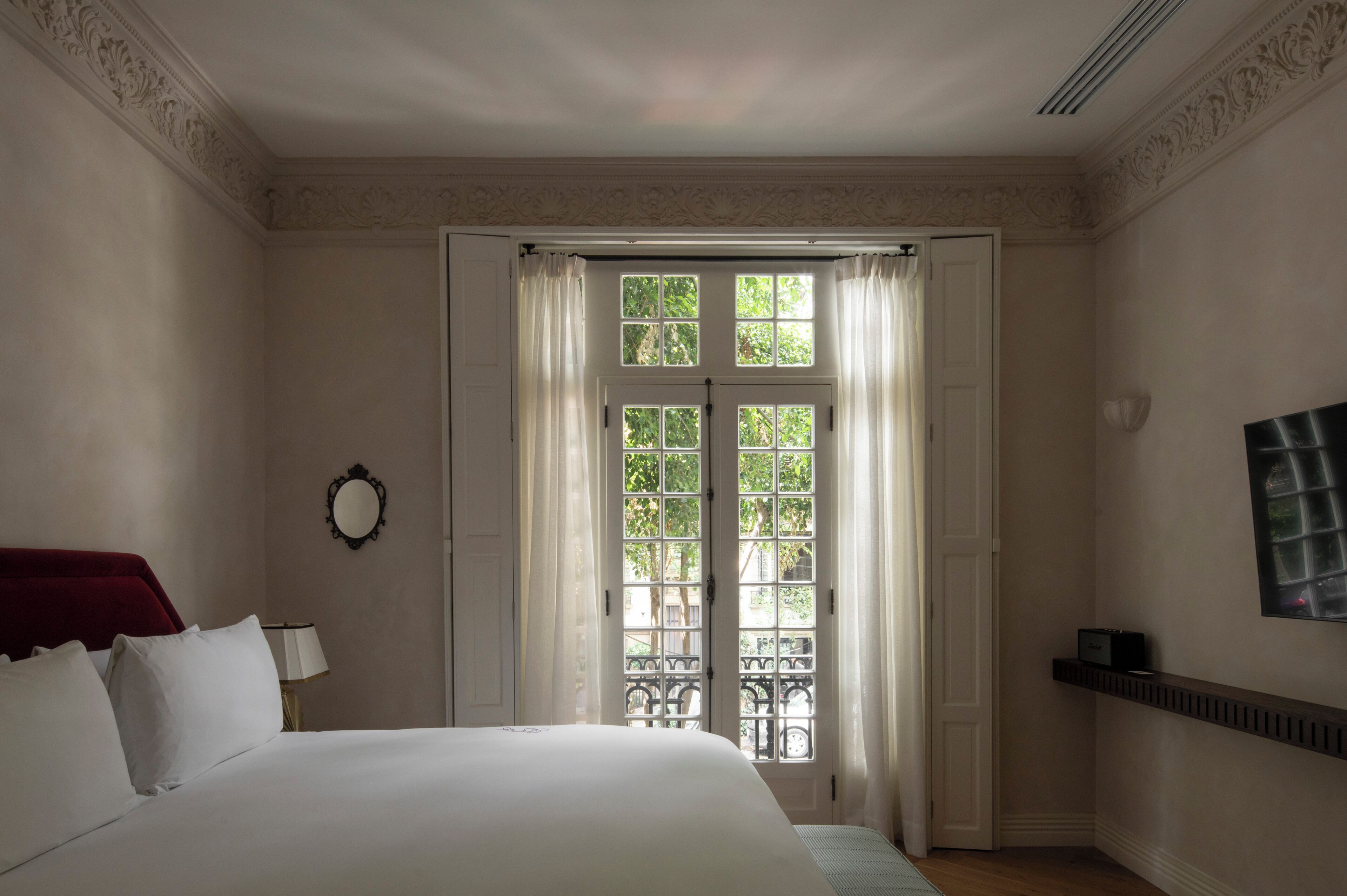Habitación de hotel con cama, espejo, ventana con cortinas y televisor de pared.