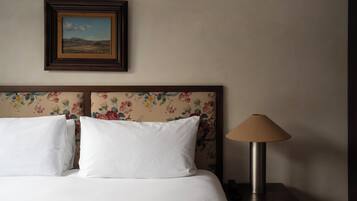 Una cama bien hecha con sábanas blancas y un cabecero floral, un cuadro de paisaje enmarcado encima, y una lámpara de noche sobre una mesita de madera.