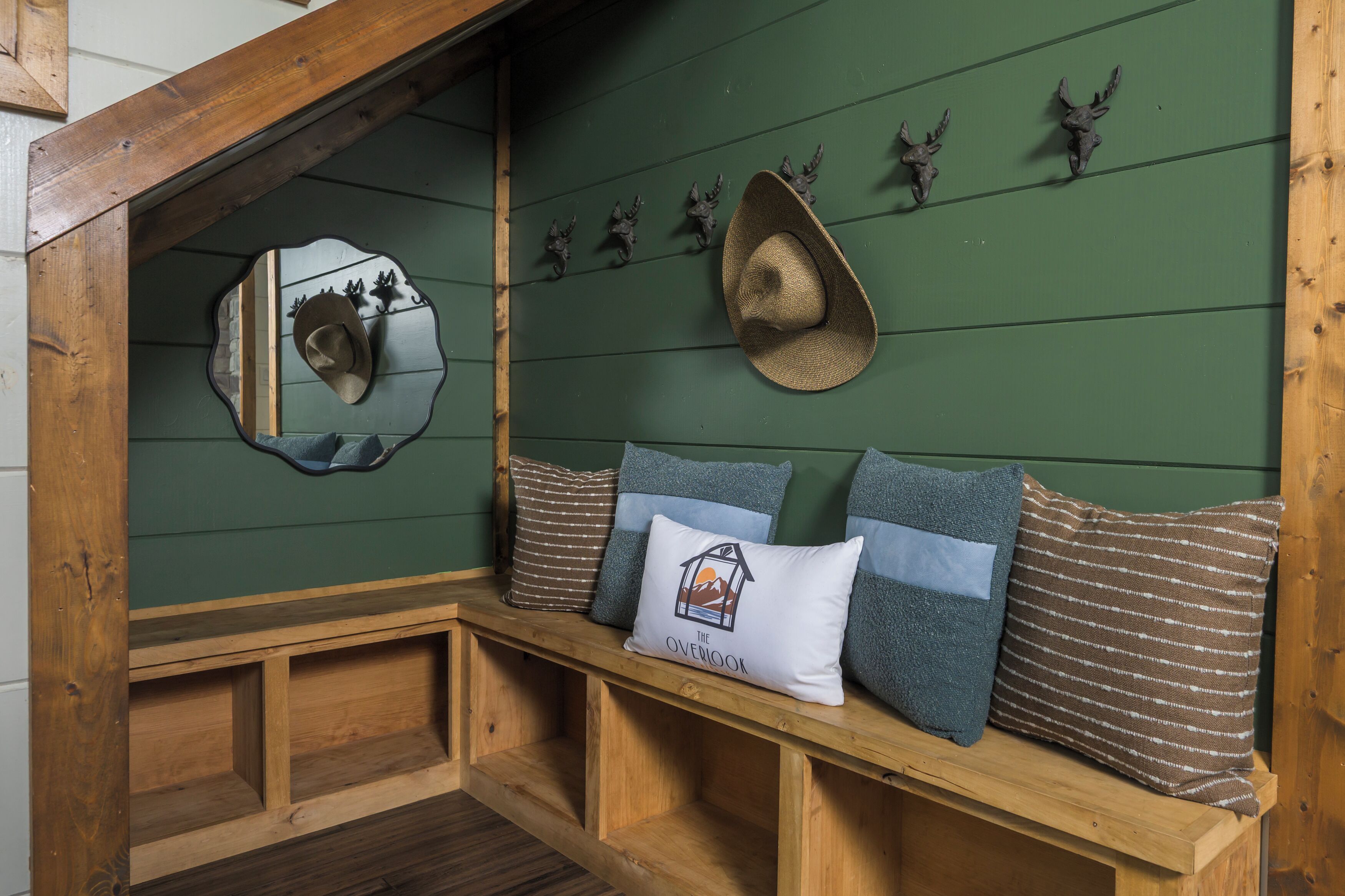 Un banco de madera con cojines, un espejo y cabezas de animales decorativas sobre una pared verde.