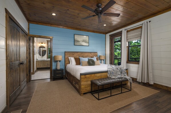 Un dormitorio con techo de madera, una cama con varias almohadas, un banco, una mesita de noche, un espejo y una ventana con cortinas.