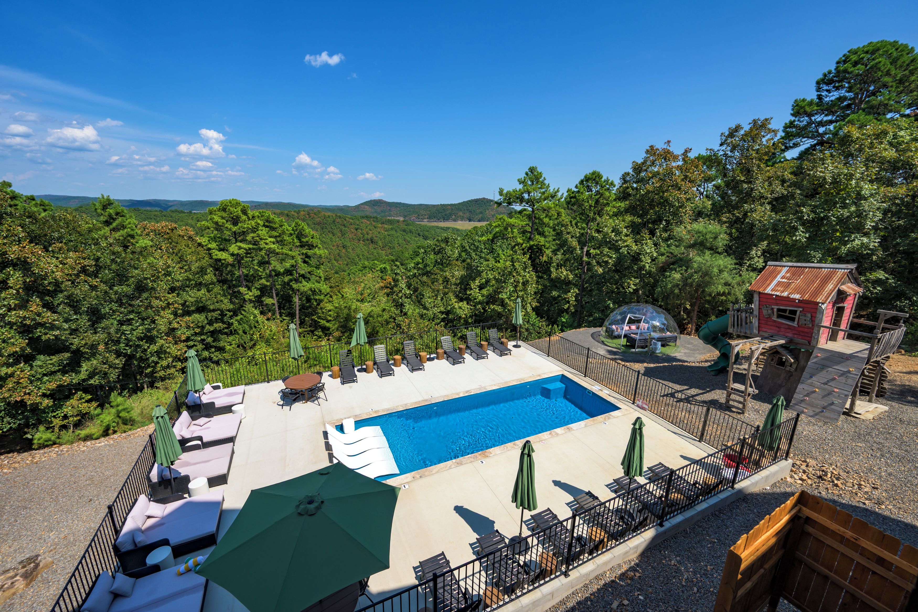 A pool area with lounge chairs and umbrellas, surrounded by a fence, with a view of a forested landscape.