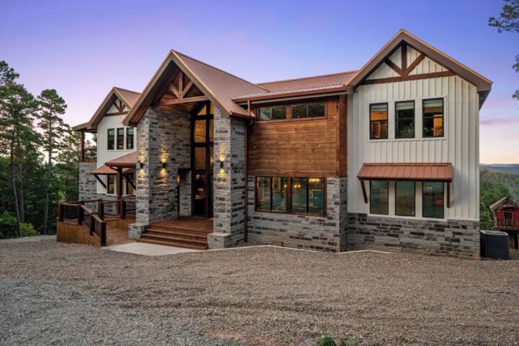 A two-story stone and wood house with a stone entrance, large windows, and a gravel driveway.
