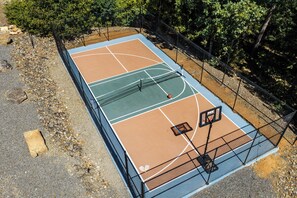 Una cancha de baloncesto al aire libre con aro, rodeada por una valla y árboles.