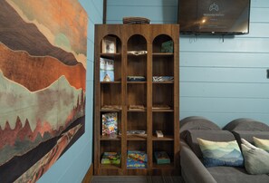 Una habitación con una estantería de madera llena de libros y juegos, un sofá con cojines y un cuadro en la pared.