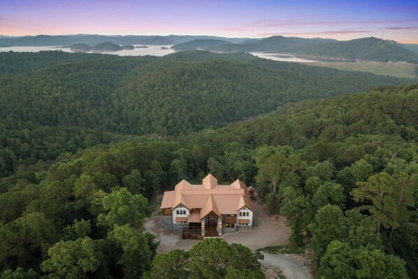 A large, two-story house with a steeply pitched roof, surrounded by dense forest.