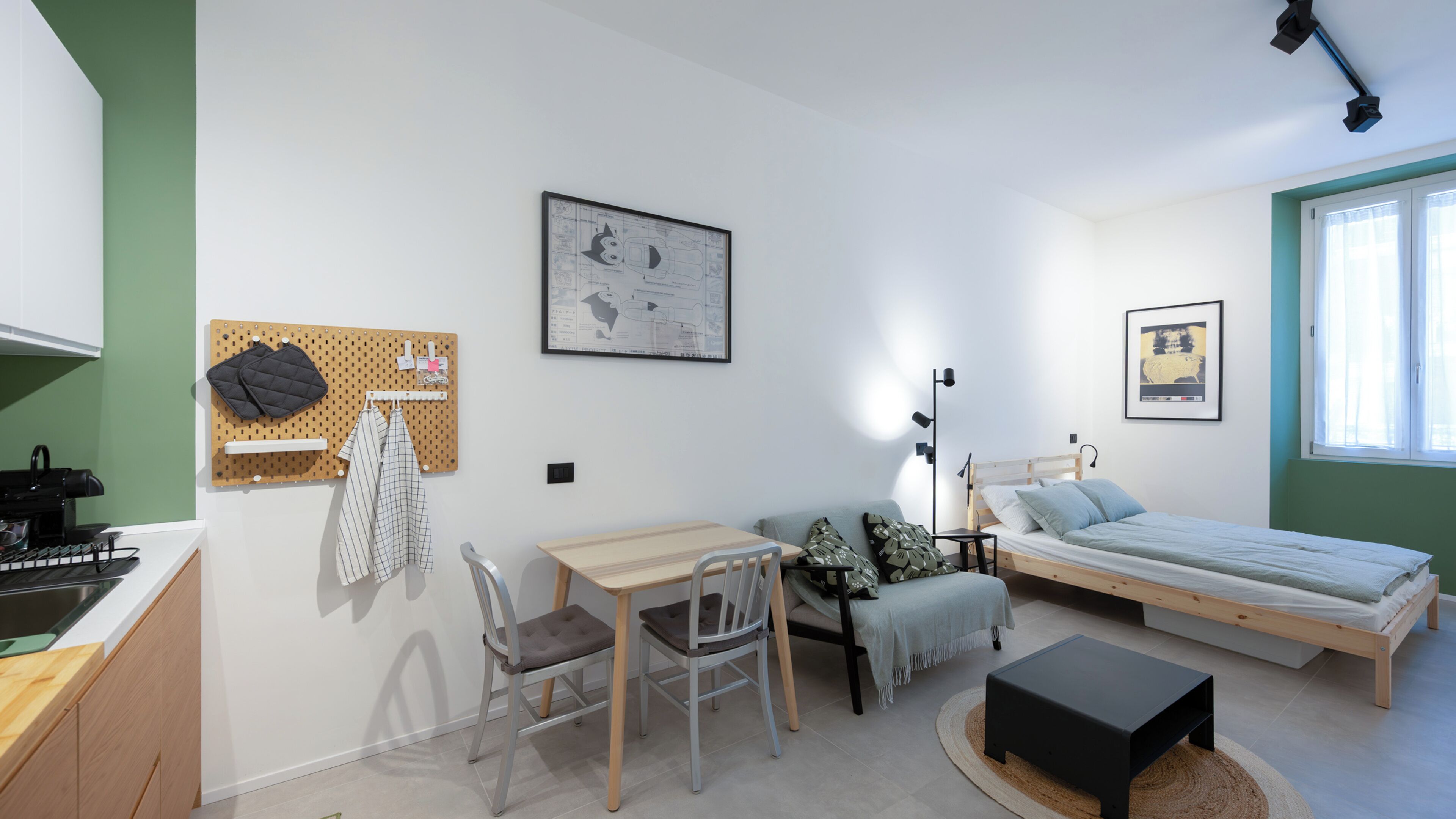 Una habitación compacta con cama, mesita de noche y escritorio con silla. Se ve un tablero de corcho con objetos en la pared, una ventana con cortinas y una lámpara en el techo.