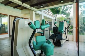 A fitness area with exercise equipment, including a leg press machine and a seated row machine, in a room with a large window and a view of greenery outside. - The Lavana Villa Gajah Jimbaran (Bali)