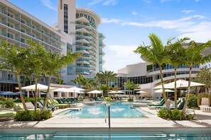 Un espace piscine d’hôtel doté de nombreuses chaises longues et de parasols, entouré de palmiers et d’immeubles modernes.