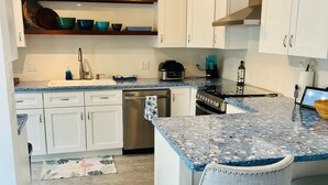 Eine Küche mit weißen Schränken, einer Granit Arbeitsplatte, einem Edelstahl-Geschirrspüler und einem eingebauten Backofen. Zwei Barhocker und ein kleiner Teppich mit tropischem Muster sind ebenfalls vorhanden.