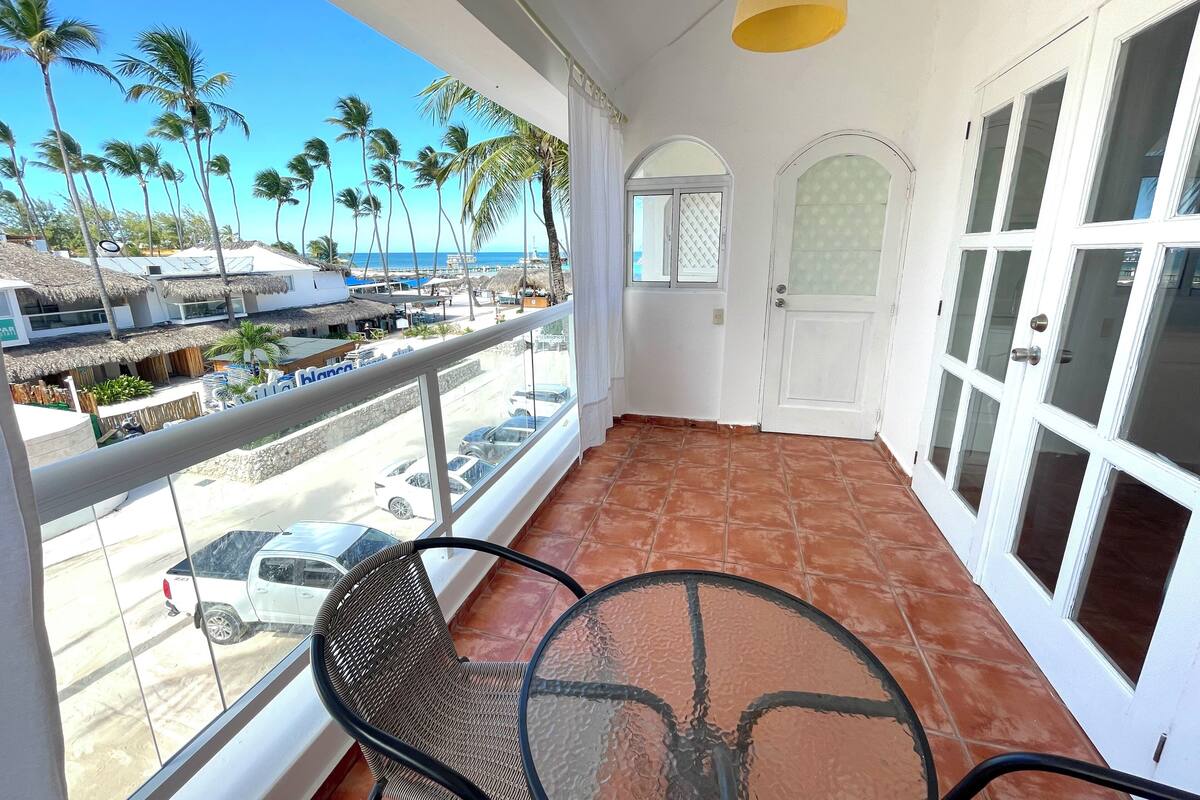 Un balcon avec une table et des chaises, une vue sur la plage et une porte dont les panneaux sont en verre dépoli.
